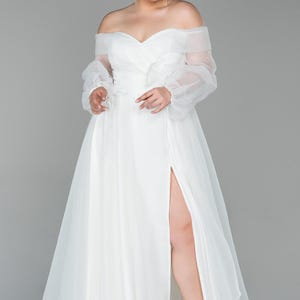 May include: A white off-the-shoulder wedding dress with sheer, long sleeves and a high slit. The dress features a flowing skirt and is paired with black heels. The model is standing in front of a grey background.