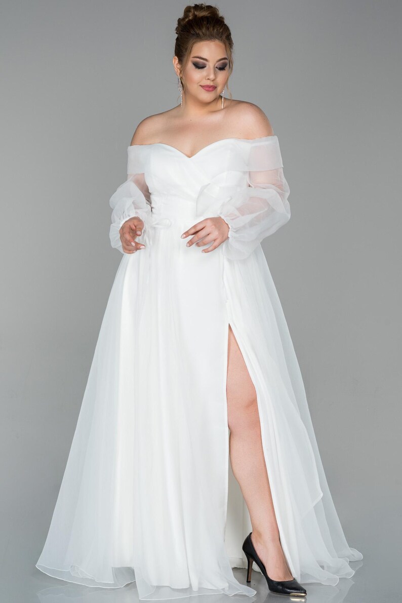 May include: A white off-the-shoulder wedding dress with sheer, long sleeves and a high slit. The dress features a flowing skirt and is paired with black heels. The model is standing in front of a grey background.
