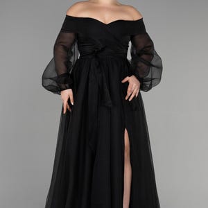 May include: A black off-the-shoulder evening gown with long, sheer sleeves and a high slit. The dress has a fitted bodice, a flowing skirt, and a tied waist. The model is wearing silver sandals.