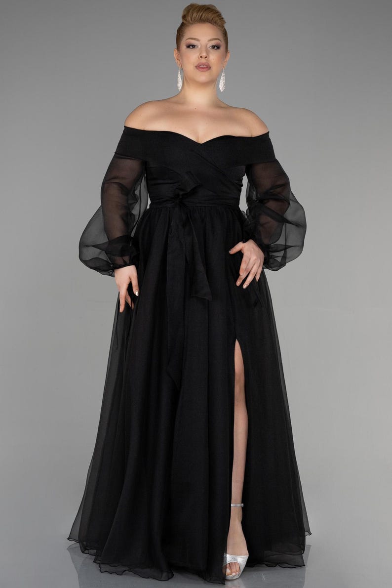 May include: A black off-the-shoulder evening gown with long, sheer sleeves and a high slit. The dress has a fitted bodice, a flowing skirt, and a tied waist. The model is wearing silver sandals.
