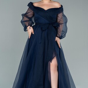 May include: A navy blue off-the-shoulder formal gown with sheer long sleeves and a thigh-high slit. The dress features a wrap-style bodice and a matching sash. The model is wearing silver heels.
