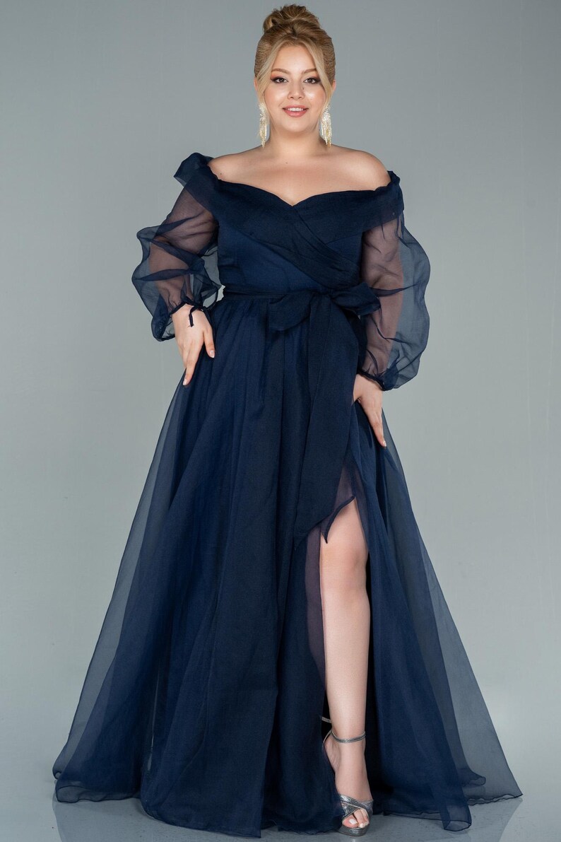 May include: A navy blue off-the-shoulder formal gown with sheer long sleeves and a thigh-high slit. The dress features a wrap-style bodice and a matching sash. The model is wearing silver heels.