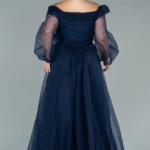 May include: A navy blue formal gown with an off-the-shoulder neckline and sheer, long sleeves. The dress features a fitted bodice and a flowing, floor-length skirt. The back of the dress has a zip closure.