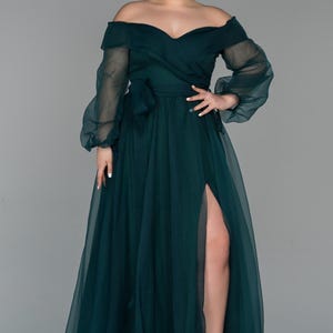 May include: A dark teal off-the-shoulder formal gown with long, sheer sleeves and a high slit. The dress features a wrap-style bodice with a bow at the waist. The model is wearing nude platform heels.