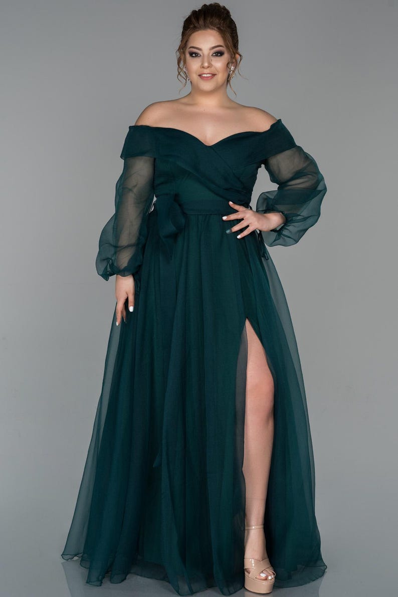 May include: A dark teal off-the-shoulder formal gown with long, sheer sleeves and a high slit. The dress features a wrap-style bodice with a bow at the waist. The model is wearing nude platform heels.