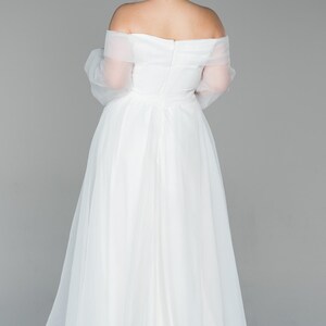 May include: A white, off-the-shoulder wedding dress with sheer sleeves and a flowing skirt. The gown has a fitted bodice and a full, floor-length skirt. The bride's hair is styled in a bun.