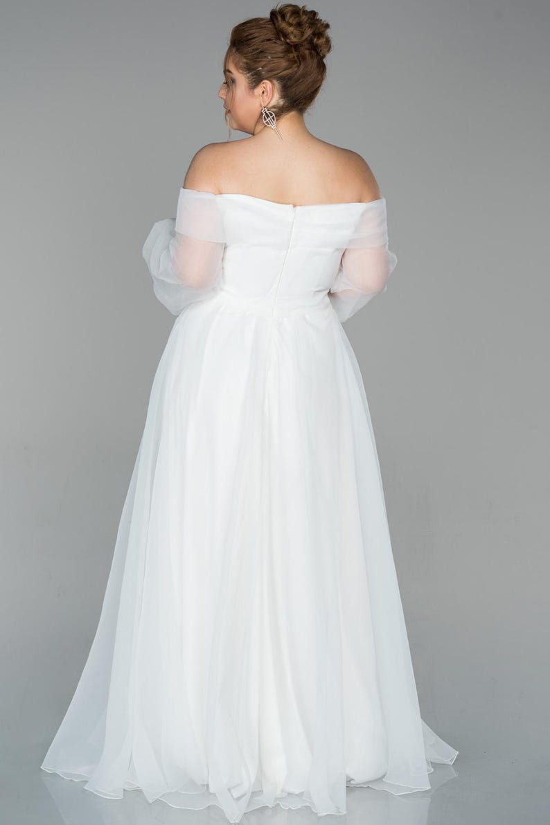 May include: A white, off-the-shoulder wedding dress with sheer sleeves and a flowing skirt. The gown has a fitted bodice and a full, floor-length skirt. The bride's hair is styled in a bun.