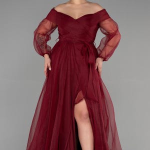 May include: A burgundy off-the-shoulder formal gown with sheer long sleeves and a thigh-high slit. The dress features a wrap-style bodice and a matching waist tie. The flowing skirt is made of a lightweight fabric.