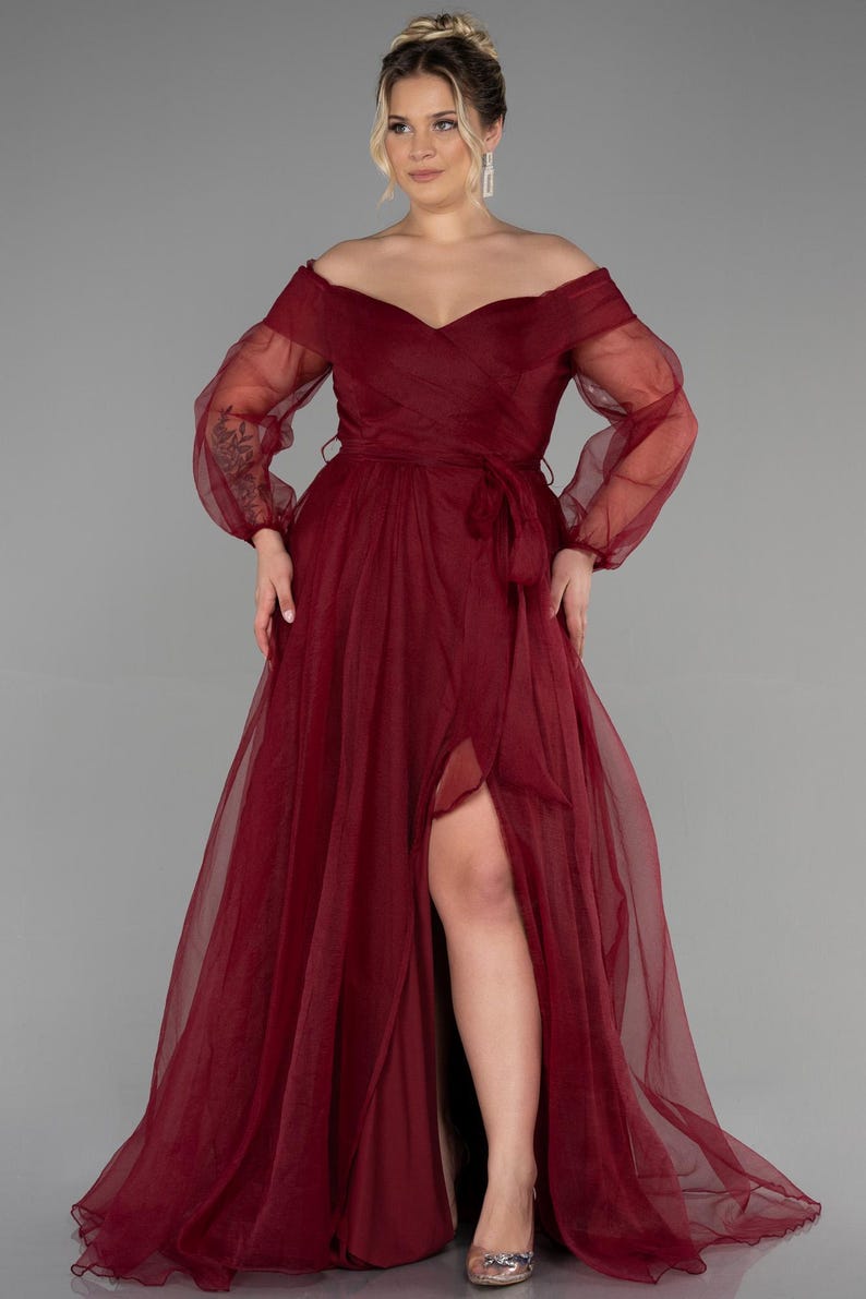 May include: A burgundy off-the-shoulder formal gown with sheer long sleeves and a thigh-high slit. The dress features a wrap-style bodice and a matching waist tie. The flowing skirt is made of a lightweight fabric.