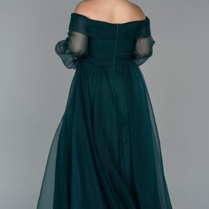 May include: A dark teal off-the-shoulder formal gown. The dress has a fitted bodice and a flowing skirt made of a sheer fabric. The sleeves are long and sheer, and the back of the dress has a zip closure. The model is facing away from the camera.