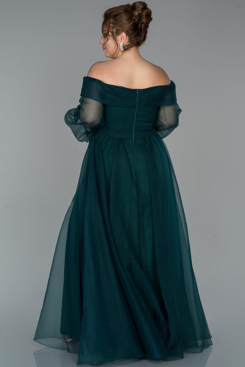 May include: A dark teal off-the-shoulder formal gown. The dress has a fitted bodice and a flowing skirt made of a sheer fabric. The sleeves are long and sheer, and the back of the dress has a zip closure. The model is facing away from the camera.