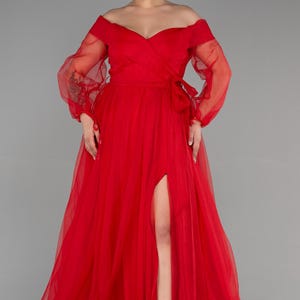 May include: A striking red off-the-shoulder gown with long, sheer sleeves. The dress has a high slit and a flowing skirt. A bow detail adorns the waist. The gown is suitable for formal events.