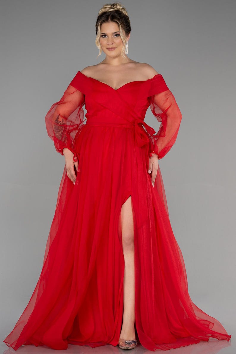 May include: A striking red off-the-shoulder gown with long, sheer sleeves. The dress has a high slit and a flowing skirt. A bow detail adorns the waist. The gown is suitable for formal events.