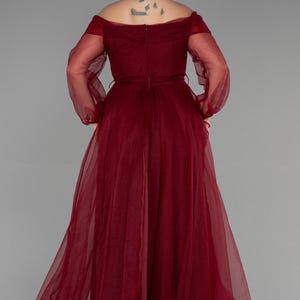 May include: A full-length, off-the-shoulder formal gown in a deep burgundy colour. The dress features long, sheer sleeves and a flowing skirt. The back of the dress has a subtle V-shape, and a thin belt cinches the waist. The fabric appears to be a lightweight chiffon.