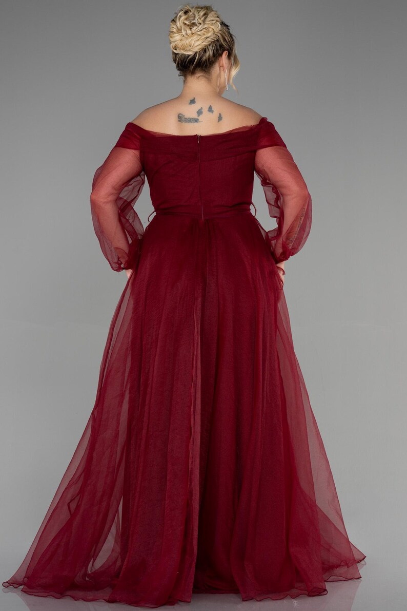 May include: A full-length, off-the-shoulder formal gown in a deep burgundy colour. The dress features long, sheer sleeves and a flowing skirt. The back of the dress has a subtle V-shape, and a thin belt cinches the waist. The fabric appears to be a lightweight chiffon.