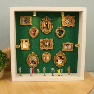 Puede incluir: Una caja de sombra blanca con un respaldo de fieltro verde exhibe marcos dorados en miniatura de varias formas. Pequeñas figuras están en la parte inferior. Una pequeña planta en maceta se encuentra sobre una mesa de madera en primer plano.