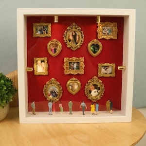 Könnte beinhalten: Eine weiße Keramiktasse mit schwarzem Henkel und dem Text "But First, Coffee" in fetter, schwarzer Schrift. Die Tasse steht vor einem verschwommenen Hintergrund, der eine gemütliche Umgebung suggeriert.