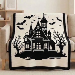 May include: A cream-colored crocheted blanket featuring a black silhouette of a haunted house, bats, and bare trees. The blanket has a black border and is draped over a beige sofa. A decorative piece for Halloween or spooky decor.