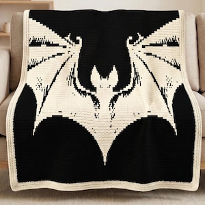 May include: A black and cream crochet blanket with a bat motif. The bat has spread wings and is centrally placed on the blanket. The blanket is draped over a beige sofa. The blanket's edges have a cream border. The blanket is approximately 150 cm x 200 cm.