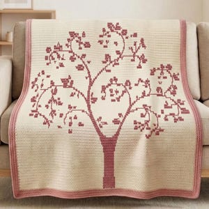 Puede incluir: Una manta de ganchillo color crema con un diseño de árbol en tonos rosas. El árbol tiene un tronco y ramas con hojas. La manta tiene un borde rosa y está colocada sobre un sofá beige.