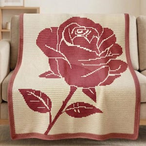 Può includere: Una coperta all'uncinetto color crema con una grande rosa pixelata nei toni del bordeaux. La coperta ha un bordo bordeaux ed è drappeggiata su un divano. Il disegno della rosa comprende uno stelo e foglie.