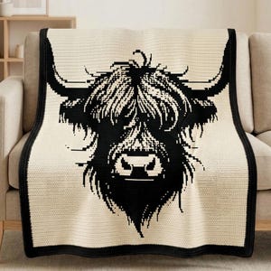 以下が含まれることがあります： ハイランド牛の顔の黒いグラフィックが特徴のクリーム色のスローブランケット。ブランケットは黒い縁取りがあり、ベージュのソファに掛けられています。牛の顔はピクセル化された線で詳細に描かれています。