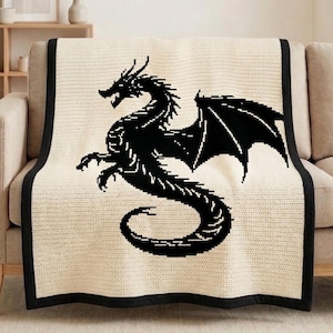 Puede incluir: Una manta crema con un diseño de dragón negro. El dragón se representa en silueta, con las alas extendidas y las garras extendidas. La manta tiene un borde negro y está sobre un sofá beige.