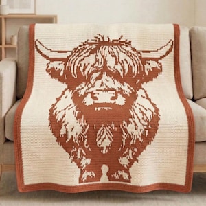 May include: A cream-colored throw blanket featuring a large, rust-colored Highland cow design. The blanket has a rust-colored border and is draped over a beige sofa. The cow's silhouette is detailed, with visible fur and horns.