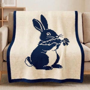 May include: A cream-colored crocheted blanket featuring a navy blue rabbit design holding a sprig of carrots. The blanket has a navy blue border and is draped over a beige sofa. The blanket is a cozy home decor item.