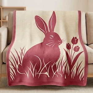 Op de afbeelding: Een gebreide deken met een bordeauxrood konijnontwerp, gras en tulpen. De deken heeft een crèmekleurige achtergrond en een bordeauxrode rand. Ideaal voor de lente of Pasen, het voegt een vleugje natuur toe aan elke woonruimte.