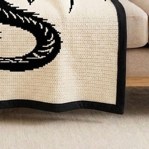May include: Cream-colored knitted throw blanket with a black border and a large black graphic design. The blanket is folded over a beige sofa, showcasing its texture and design. The blanket is made of a soft, knitted material.