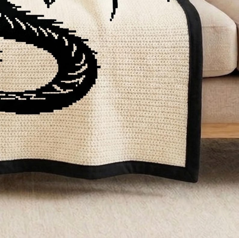 May include: Cream-colored knitted throw blanket with a black border and a large black graphic design. The blanket is folded over a beige sofa, showcasing its texture and design. The blanket is made of a soft, knitted material.