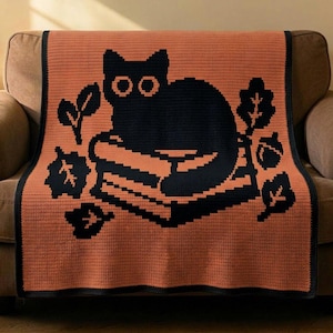 Pode incluir: Uma manta de crochê retangular com fundo laranja queimado e um design gráfico preto. O desenho retrata um gato preto sentado sobre uma pilha de livros, cercado por várias formas de folhas e uma bolota, tudo contornado por uma borda preta.