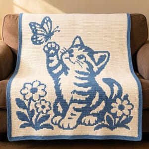 May include: A rectangular crochet blanket featuring a cream-colored background with a blue graphic pattern. The design depicts a kitten reaching toward a butterfly, surrounded by two flowers. The blanket is finished with a solid blue border.