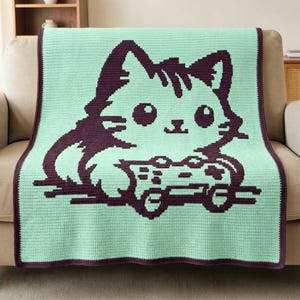 Peut inclure: Une couverture en crochet rectangulaire avec un fond vert menthe et un motif pixelisé violet foncé représentant un chat tenant une manette de jeu vidéo. La couverture présente une bordure violet foncé qui encadre tout le design.