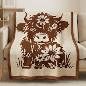 Puede incluir: Una manta de crochet color crema con un diseño de vaca de las Tierras Altas marrón adornada con flores. La manta tiene un borde marrón y está colocada sobre un sofá beige. El diseño de la vaca incluye detalles florales y hierba.