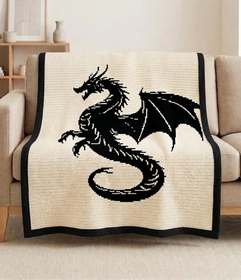 May include: A cream-colored throw blanket with a black dragon design. The dragon is depicted in a stylized silhouette, with wings spread and claws extended. The blanket has a black border and is draped over a beige sofa.