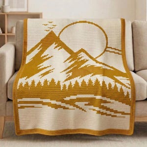 Puede incluir: Una manta crema con un diseño de montaña y sol amarillo mostaza. La manta presenta un paisaje con montañas, árboles y un gran sol. Los bordes están ribeteados con un borde amarillo mostaza. La manta está sobre un sofá beige.