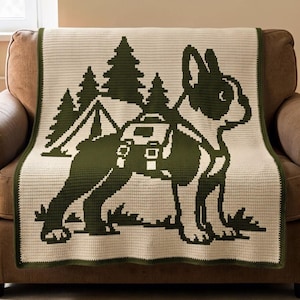May include: A rectangular crochet blanket featuring a two-tone design in cream and dark forest green. The graphic depicts a dog wearing a backpack standing in front of a tent and pine trees. The blanket has a solid dark green border edge.