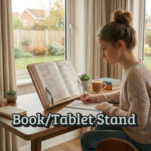 May include: A wooden book and tablet stand holds an open book with handwritten notes. A person is writing in a notebook at a desk. The stand has metal clips to hold the book open. The text "Book/Tablet Stand" is at the bottom.