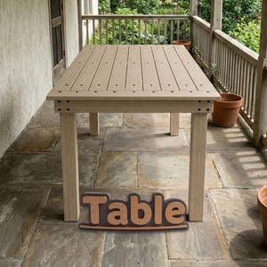Könnte beinhalten: Ein rechteckiger hellbrauner Tisch mit einer Lattenoberfläche, bei der die Schraubenköpfe auf jeder Planke sichtbar sind. Der Tisch hat vier gerade Beine und einen stabilen Rahmen. Ein dekoratives Schild mit dem Wort Table steht davor.