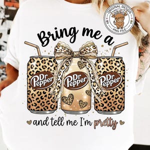 May include: White t-shirt featuring a graphic of three Dr. Pepper cans with leopard print and a bow. The text reads "Bring me a Dr. Pepper and tell me I'm pretty."