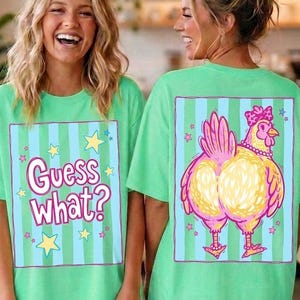 May include: Two women wearing matching green t-shirts. The front of the shirt has the text "Guess What?" inside a blue and green striped box with stars. The back of the shirt features a cartoon chicken design.