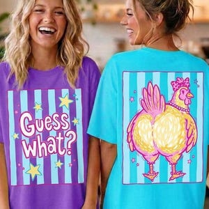May include: Two women wearing oversized t-shirts. One shirt is purple with the text "Guess What?" and vertical blue stripes. The other is turquoise with a cartoon chicken design on the back. Both shirts have a relaxed fit.
