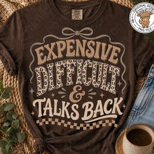 Puede incluir: Una camiseta de color marrón oscuro con el texto "EXPENSIVE DIFFICULT & TALKS BACK" en letras de color crema y estampado de leopardo. El diseño incluye un lazo decorativo en la parte superior y un patrón de cuadros en la parte inferior.