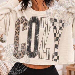 May include: A cream-colored cropped sweatshirt featuring the word COZY in large block letters. The letter C has a leopard print, the O has a distressed black texture, the Z has vertical black stripes, and the Y has a black and white checkerboard pattern.