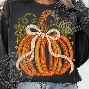 May include: A charcoal grey crewneck sweatshirt featuring a large graphic of an orange pumpkin with a beige ribbon bow tied around its stem. The pumpkin is surrounded by green leaves, curly vines, and small orange star accents on the front.
