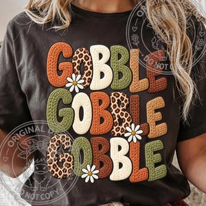 May include: A dark gray t-shirt featuring the text GOBBLE GOBBLE GOBBLE in a stacked design. The letters have a textured knit appearance in orange, cream, olive green, brown, and leopard print, accented with small white daisy flower graphics.