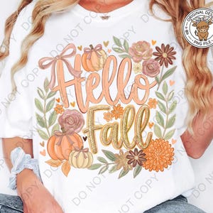 Puede incluir: Una camiseta blanca con un estampado gráfico que incluye el texto Hello Fall en letras color melocotón y dorado. El diseño presenta calabazas naranjas, un lazo rosa y varias flores de colores otoñales y hojas verdes decorativas.