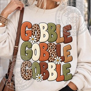 May include: A cream-colored crewneck sweatshirt featuring the text GOBBLE GOBBLE GOBBLE in a stacked design. The letters have a textured knit appearance in shades of orange, brown, green, and leopard print, accented with small white daisy flowers.